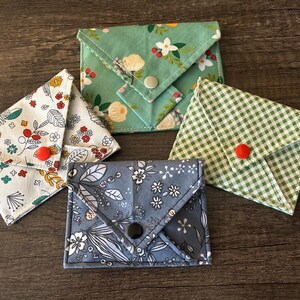 May include: An assortment of small, fabric wallets in various floral and patterned designs. The wallets are in shades of green, blue, and white, with red and black button closures. The wallets are arranged on a dark wooden surface.