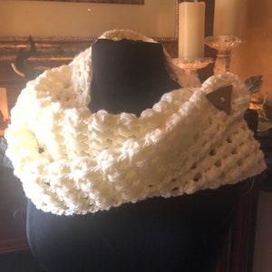 May include: A white crocheted infinity scarf with a woven texture. The scarf is displayed on a black mannequin.