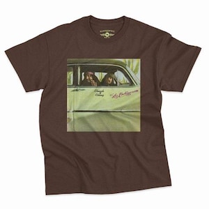 Könnte beinhalten: Ein dunkelbraunes Kurzarm-T-Shirt mit einem quadratischen Grafikdruck von zwei Personen in einem grünen Auto. Der Text "Cheech & Chong" und "Los Cochinos" ist in einer Schreibschrift auf der Autotür innerhalb des Grafikdesigns abgedruckt.