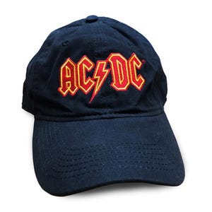 May include: A navy blue baseball cap featuring the AC/DC logo embroidered on the front. The logo consists of red lettering with a yellow outline, including a central lightning bolt symbol. The cap has a curved brim and stitched detailing.