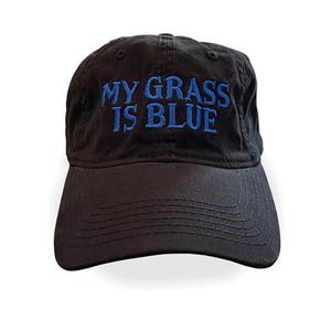 May include: A black baseball cap featuring the embroidered text MY GRASS IS BLUE in a bright blue font. The cap has a structured crown, a curved brim with visible stitching, and eyelets on the top panels for ventilation and a classic design.