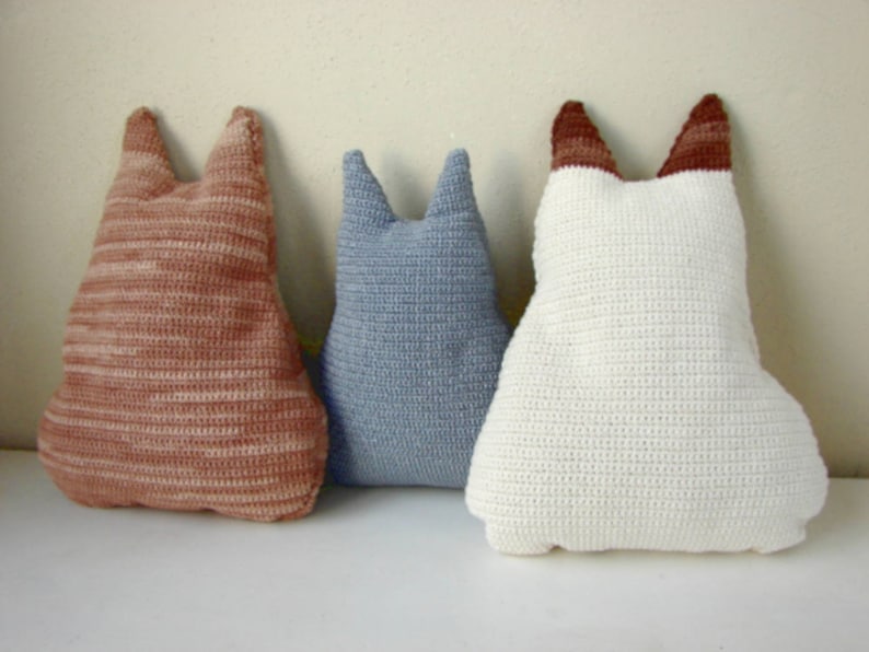 May include: Three crocheted cat-shaped pillows in different colors: brown and white, blue, and white. The pillows are arranged in a row on a white surface.