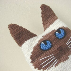 May include: Crocheted Siamese cat toy with brown and white fur and blue eyes. The cat has a white nose and whiskers.