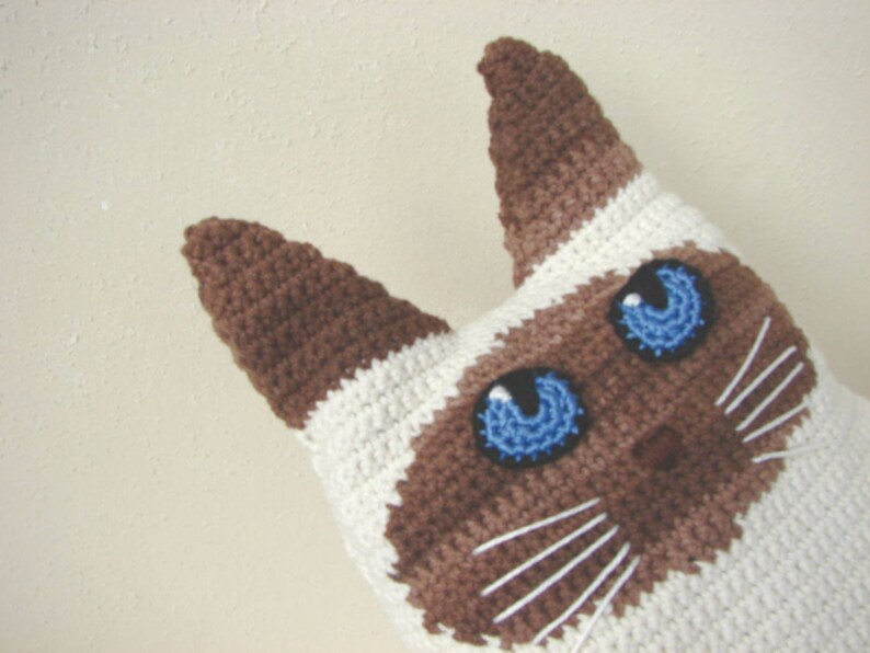 May include: Crocheted Siamese cat toy with brown and white fur and blue eyes. The cat has a white nose and whiskers.