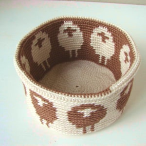 May include: A crocheted basket with a brown and white pattern of sheep. The basket is round and has a wide opening.