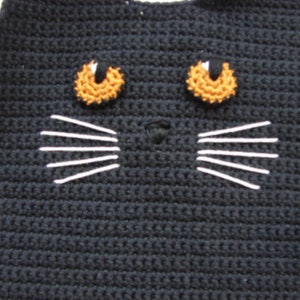 May include: Close-up of a black crocheted cat face with orange eyes and white whiskers.