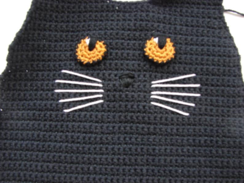 May include: Close-up of a black crocheted cat face with orange eyes and white whiskers.