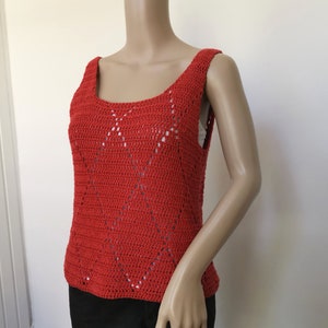 May include: A sleeveless, red crochet top with a scoop neckline and diamond-shaped openwork design. The top is made of a textured yarn and appears to be handmade. The garment is displayed on a mannequin.