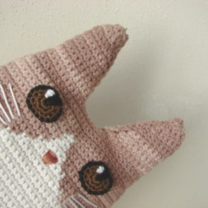 May include: A crocheted cat toy with brown eyes and a pink nose. The cat is made with white and pink yarn.