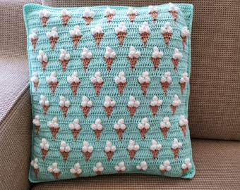 May include: A turquoise blue crocheted pillow with a repeating pattern of brown ice cream cones and white ice cream scoops.