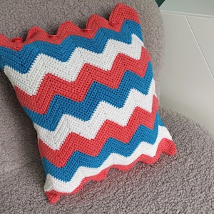 May include: A crocheted pillow with a chevron pattern in shades of red, white, and blue. The pillow is sitting on a gray, fuzzy surface.