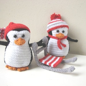Puede incluir: Dos juguetes de pingüino de crochet, uno con un gorro a rayas rojas y blancas y una bufanda roja, el otro con un gorro rojo con un pompón blanco. El pingüino con el gorro a rayas está sentado en un trineo gris con rayas rojas y blancas.
