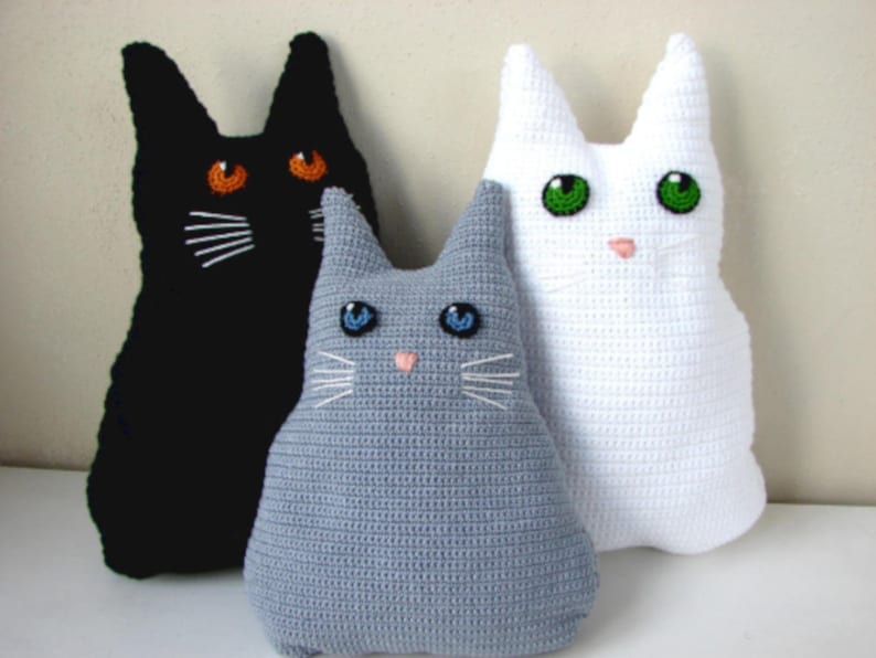 May include: Three crocheted cat plushies, one black, one gray, and one white. Each plush has embroidered facial features, including eyes, a nose, and whiskers.