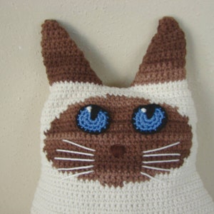 May include: A crocheted Siamese cat pillow with brown ears and a white body. The cat has blue eyes and a brown nose.