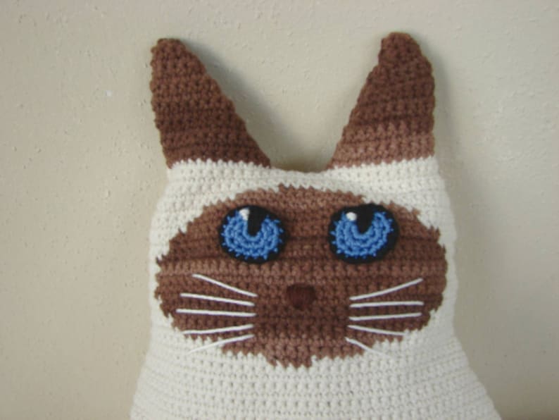 May include: A crocheted Siamese cat pillow with brown ears and a white body. The cat has blue eyes and a brown nose.