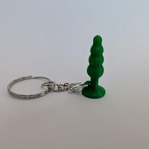 May include: A green, 3D-printed keychain charm shaped like a stylized tree. The charm is attached to a silver keyring with a small chain and clasp. The charm has a textured, layered design.