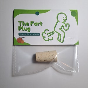 Könnte beinhalten: Ein weißer Korken mit einer grün-weißen Karte, auf der "The Fart Plug" und "Stops pesky fart attacks!" steht. Die Karte zeigt eine Karikatur einer Person mit einer Furzkwolke dahinter.