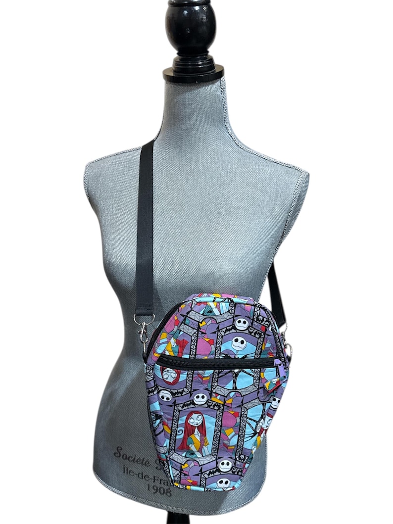 May include: A coffin-shaped crossbody bag with a black strap, featuring a colourful print of characters and scenes. The bag has a black zip and is displayed on a mannequin.