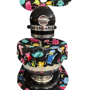 May include: A black KitchenAid stand mixer featuring a fabric cover set with a black background and a colorful pattern of various teapots. The set includes a bowl cover with an elastic edge and a matching decorative bow attached to the top head.
