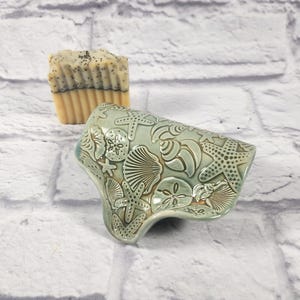 May include: A seafoam green ceramic soap dish with embossed starfish, seashells, and sand dollars. A bar of soap with a light brown and dark brown striped design is in the background. The soap dish is approximately 6 inches long.