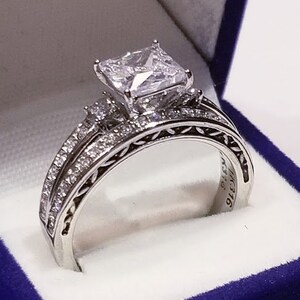 Puede incluir: Un anillo de compromiso de plata con un gran diamante de talla coj&iacute;n. El anillo est&aacute; engastado en una banda delicada e intrincada con diamantes m&aacute;s peque&ntilde;os. El anillo est&aacute; en una caja de terciopelo azul.