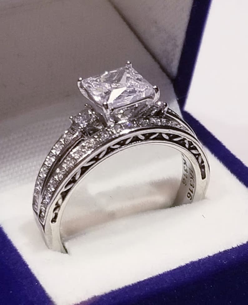 Puede incluir: Un anillo de compromiso de plata con un gran diamante de talla coj&iacute;n. El anillo est&aacute; engastado en una banda delicada e intrincada con diamantes m&aacute;s peque&ntilde;os. El anillo est&aacute; en una caja de terciopelo azul.
