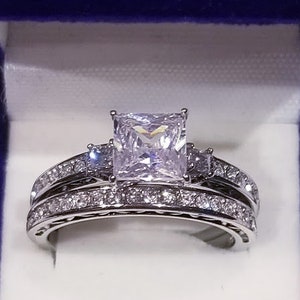 Puede incluir: Un anillo de plata con una piedra grande de circonita c&uacute;bica de corte cuadrado rodeada de piedras m&aacute;s peque&ntilde;as. El anillo est&aacute; sobre un fondo blanco.