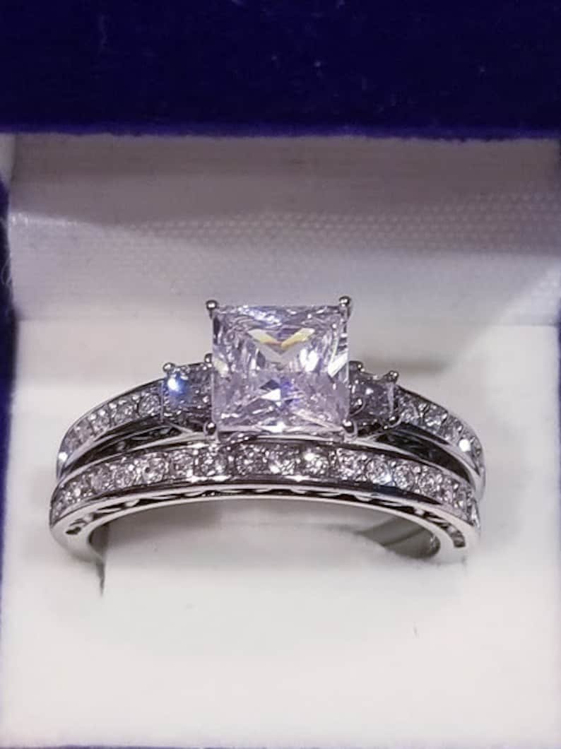 Puede incluir: Un anillo de plata con una piedra grande de circonita c&uacute;bica de corte cuadrado rodeada de piedras m&aacute;s peque&ntilde;as. El anillo est&aacute; sobre un fondo blanco.