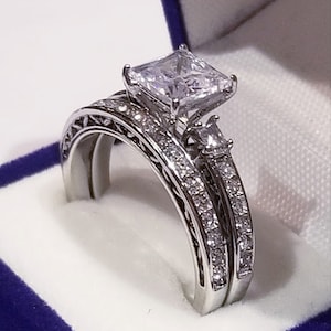 Puede incluir: Un juego de anillos de boda de plata con un gran diamante de talla cuadrada y peque&ntilde;os diamantes de acento. Los anillos se muestran en una caja de terciopelo azul.