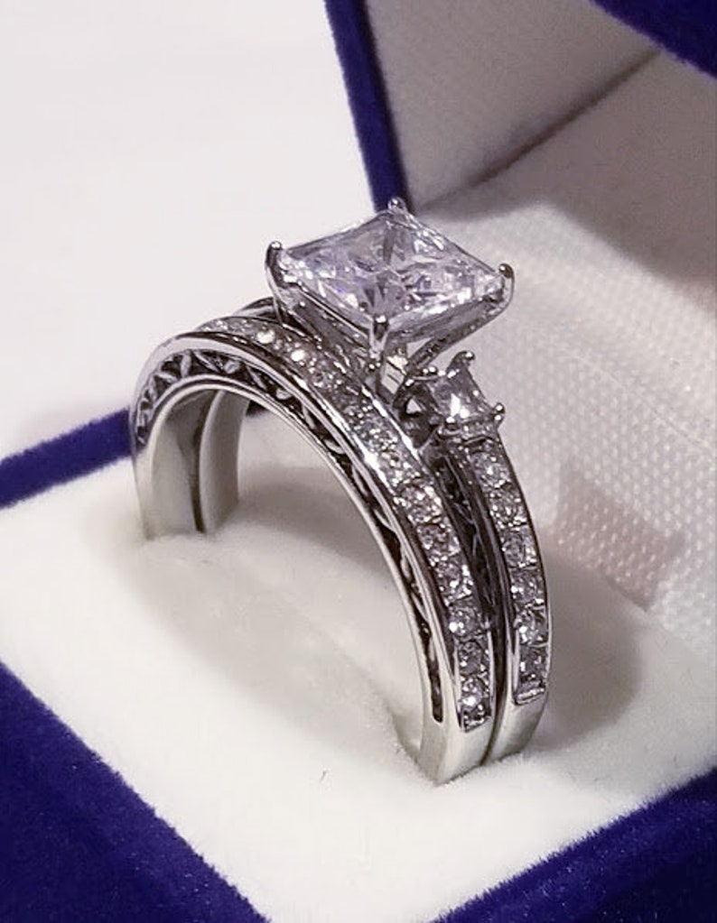 Puede incluir: Un juego de anillos de boda de plata con un gran diamante de talla cuadrada y peque&ntilde;os diamantes de acento. Los anillos se muestran en una caja de terciopelo azul.