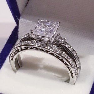 Puede incluir: Un anillo de compromiso de plata con un gran diamante central de talla cuadrada y un diamante m&aacute;s peque&ntilde;o en la banda. El anillo est&aacute; engastado en un dise&ntilde;o delicado y ornamentado.
