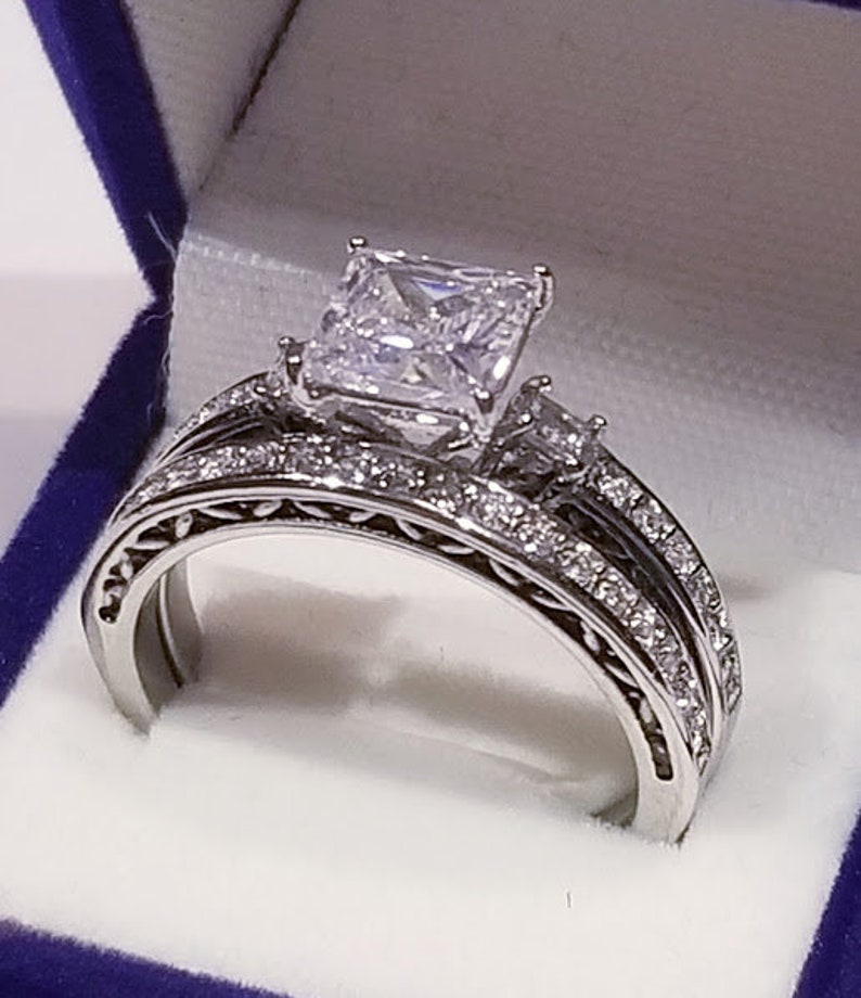 Puede incluir: Un anillo de compromiso de plata con un gran diamante central de talla cuadrada y un diamante m&aacute;s peque&ntilde;o en la banda. El anillo est&aacute; engastado en un dise&ntilde;o delicado y ornamentado.