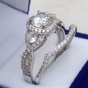 May include: A silver engagement ring set with a large round center stone surrounded by smaller stones. The band is twisted and features a smaller pear-shaped stone on each side of the center stone.
