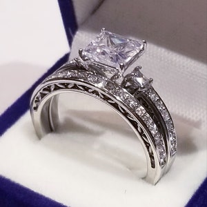Puede incluir: Un anillo de compromiso de plata con un gran diamante central de talla cuadrada y una banda a juego con diamantes m&aacute;s peque&ntilde;os.