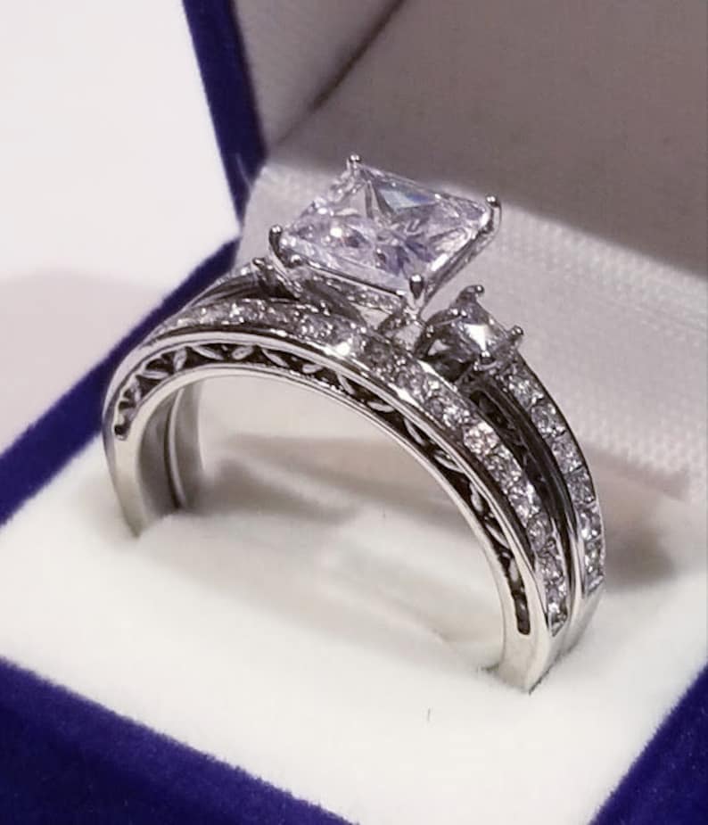 Puede incluir: Un anillo de compromiso de plata con un gran diamante central de talla cuadrada y una banda a juego con diamantes m&aacute;s peque&ntilde;os.