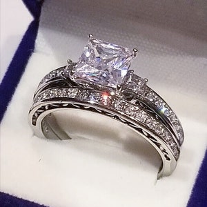 May include: A silver wedding ring set with a large, square-cut diamond center stone. The band is decorated with smaller diamonds.
