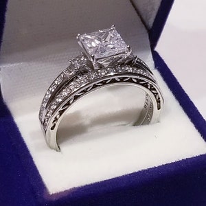 Puede incluir: Un anillo de compromiso de plata con un gran diamante de talla coj&iacute;n y diamantes m&aacute;s peque&ntilde;os en la banda. El anillo est&aacute; en una caja de anillo de terciopelo azul.