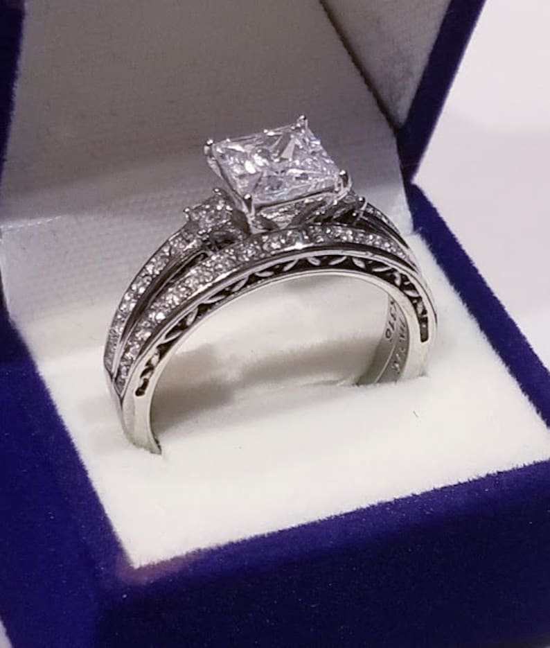 Puede incluir: Un anillo de compromiso de plata con un gran diamante de talla coj&iacute;n y diamantes m&aacute;s peque&ntilde;os en la banda. El anillo est&aacute; en una caja de anillo de terciopelo azul.