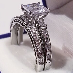 Puede incluir: Un anillo de compromiso de plata con una gran piedra preciosa cuadrada transparente. El anillo est&aacute; engastado con piedras preciosas transparentes m&aacute;s peque&ntilde;as alrededor de la piedra principal y en la banda.
