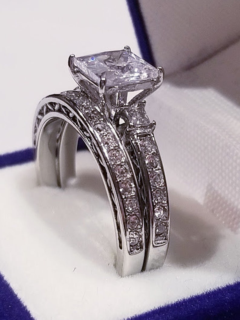 Puede incluir: Un anillo de compromiso de plata con una gran piedra preciosa cuadrada transparente. El anillo est&aacute; engastado con piedras preciosas transparentes m&aacute;s peque&ntilde;as alrededor de la piedra principal y en la banda.
