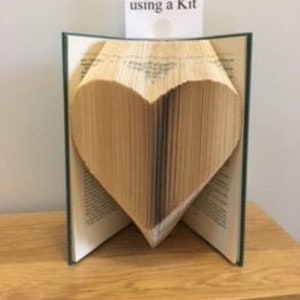 May include: A book folded into the shape of a heart. The book is open and the pages are folded to create a three-dimensional heart shape. The book is on a wooden surface. A small white card is attached to the book with the text "Fold me using a Kit".