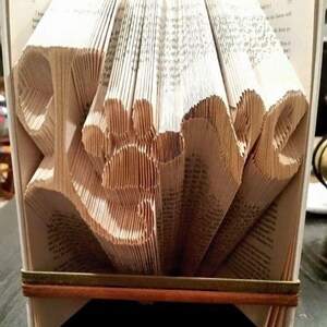 Peut inclure: Un livre plié en forme du mot "Hope". Le livre est fait de papier blanc avec du texte noir. Le livre est posé sur une surface brun foncé.