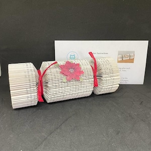 May include: A red and white paper Christmas cracker made from a book with a red poinsettia flower decoration. The cracker is tied with red ribbon and has instructions for making it.
