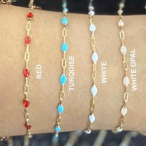 May include: Six gold-tone chain bracelets featuring small oval-shaped beads in pink, red, turquoise, white, white opal, and white pearlescent. Each bracelet is labeled with its respective color name: PINK, RED, TURQOISE, WHITE, WHITE OPAL, WHITE PEARLESCENT.