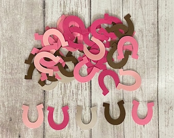 May include: A pile of pink and brown paper cutouts shaped like horseshoes. The horseshoes are scattered on a light wood background.