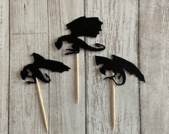 May include: Three black dragon-shaped cupcake toppers on wooden sticks. The toppers are made of a dark material and are arranged against a light wood-grain background. The dragons have detailed wings and tails.