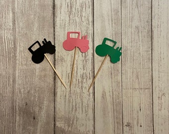 May include: Three tractor cupcake toppers, one black, one pink, and one green. The toppers are made of cardstock and are attached to wooden skewers.