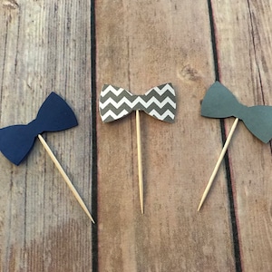 May include: Three bow tie cupcake toppers. One is navy blue, one is gray with a white chevron pattern, and one is solid gray.