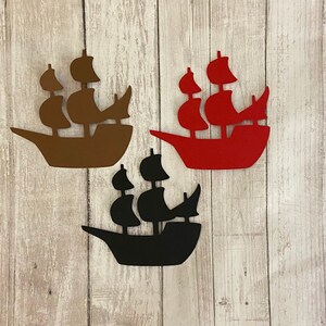 Puede incluir: Tres recortes de papel de barcos en diferentes colores: marrón, rojo y negro. Los barcos tienen todos el mismo diseño, con tres velas y una proa puntiaguda.