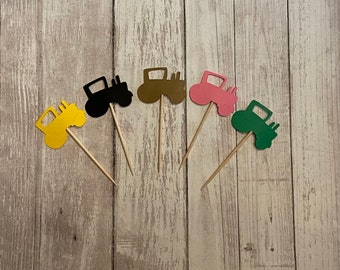 May include: Five tractor cupcake toppers in different colors: yellow, black, brown, pink, and green. The toppers are made of paper and have a stick attached to the back.